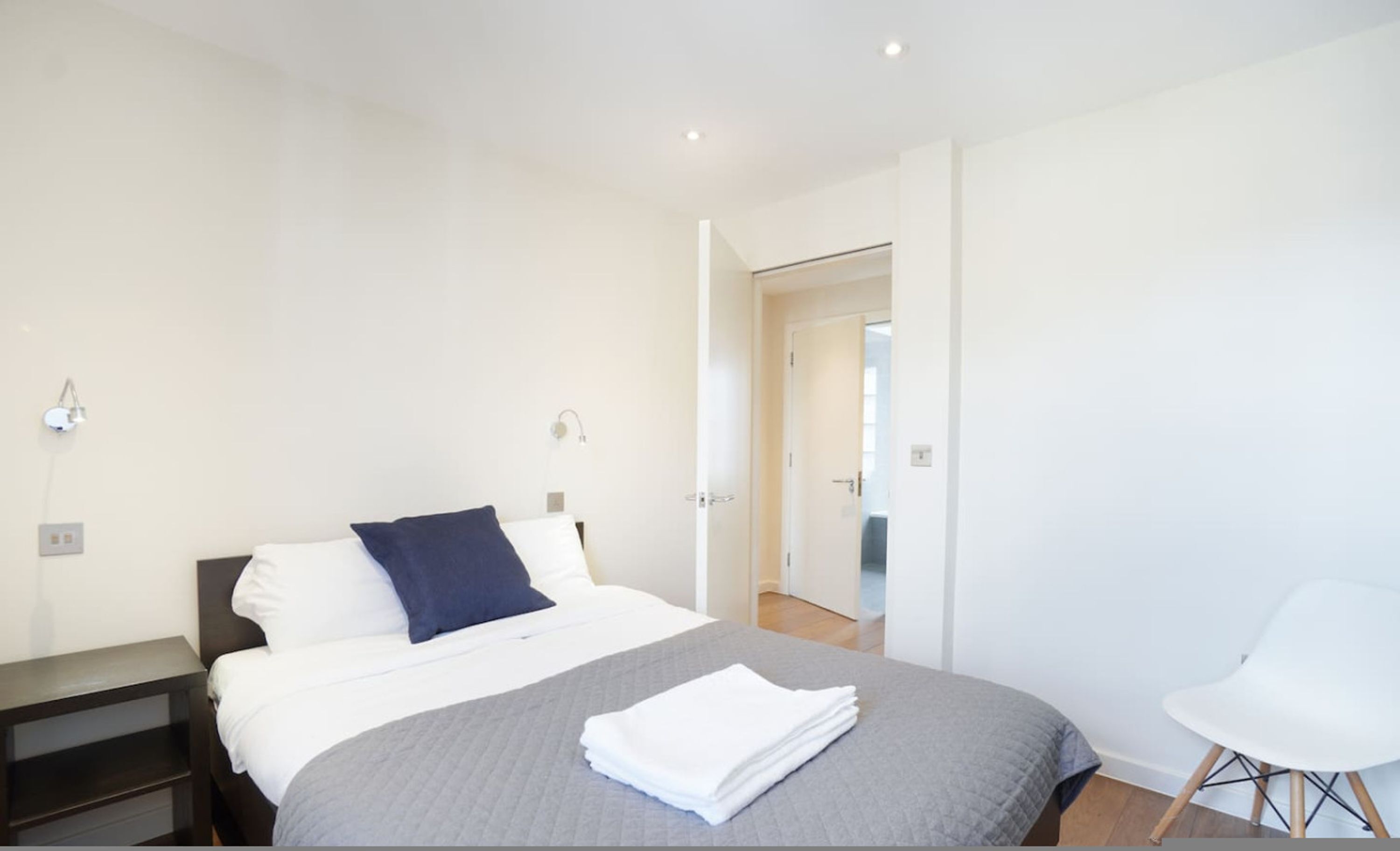 Un dormitorio moderno con cama, mesitas de noche, una silla y una puerta que da a otra habitación.