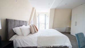 A bedroom with a bed, a nightstand, a chair, a wardrobe, and a window with a view of a building. - Stylish 3 Bedroom Flat CRC (London)