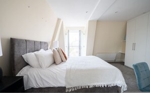 A bedroom with a bed, a nightstand, a chair, a wardrobe, and a window with a view of a building. - Stylish 3 Bedroom Flat CRC (London)