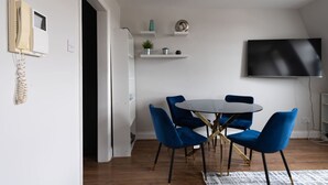 Een moderne eethoek met een ronde tafel en blauwe stoelen, een flatscreen televisie aan de wand en een klein kastje met decoratieve objecten.
