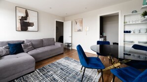 Een moderne woonkamer met een grijze bank, blauwe stoelen, een zwarte eettafel en een kamerkleed met een patroon.