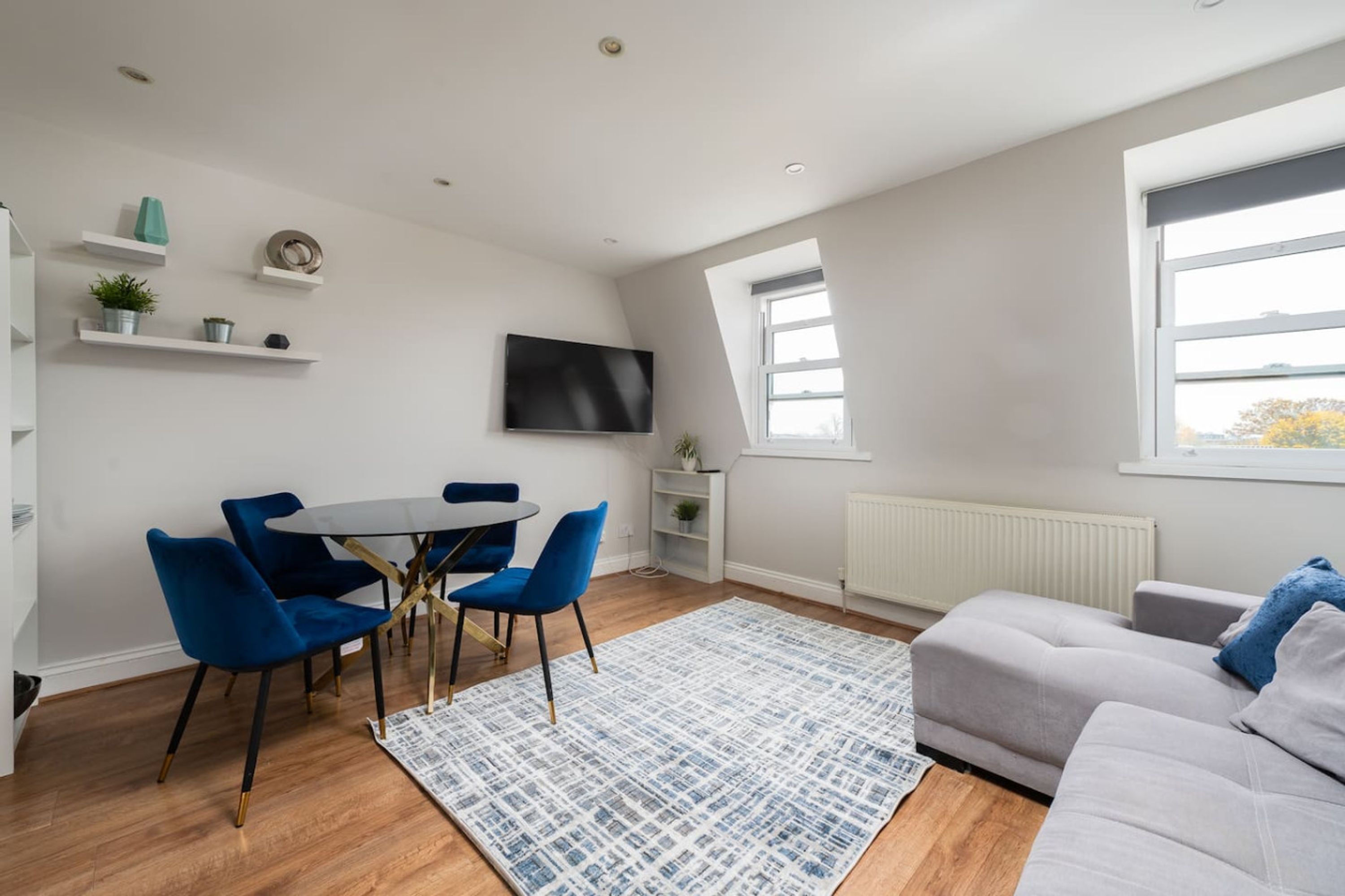 Un salon moderne avec un canapé gris, des fauteuils bleus, une table ronde et un téléviseur fixé au mur.