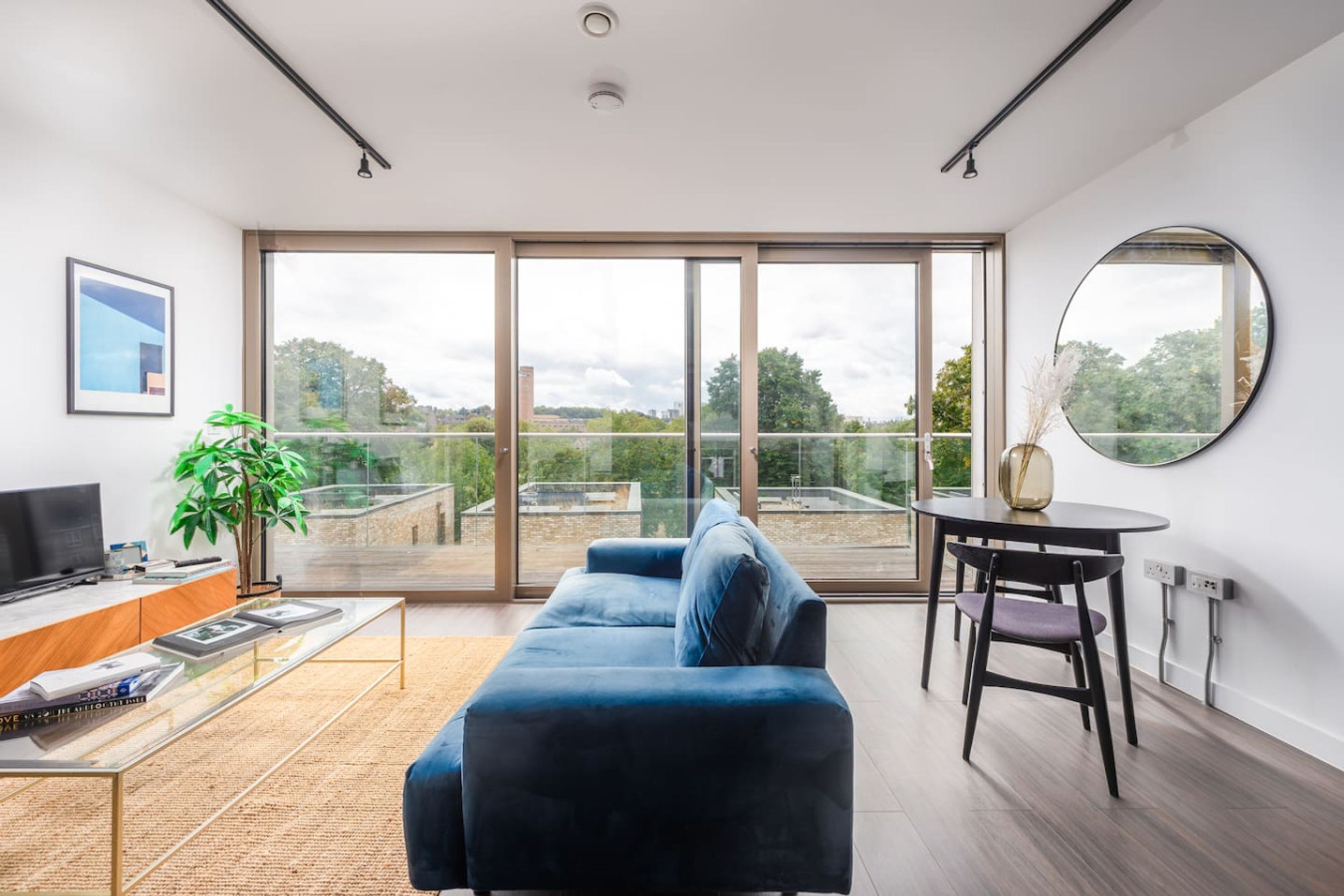 Un salon moderne doté d’une grande porte-fenêtre coulissante donnant sur un balcon, d’un canapé bleu, d’une petite table ronde et d’un téléviseur posé sur un meuble en bois.