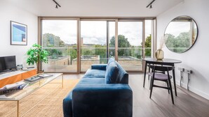 Un soggiorno moderno con una grande porta-finestra che si apre su un balcone, un divano blu, un piccolo tavolino rotondo e un televisore su un mobile in legno.