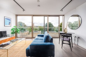 Ein modernes Wohnzimmer mit einer großen Glasschiebetür, die auf einen Balkon führt, einem blauen Sofa, einem kleinen runden Tisch und einem Fernseher auf einem Holzgestell.