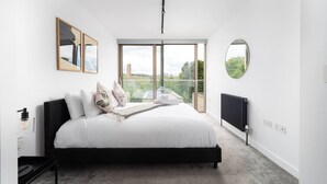 Camera da letto moderna con un letto grande, vista su vegetazione e edifici, e uno specchio rotondo appeso al muro.