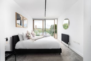 Ein modernes Schlafzimmer mit einem großen Bett, Blick auf Grünflächen und Gebäude sowie einem runden Spiegel an der Wand.