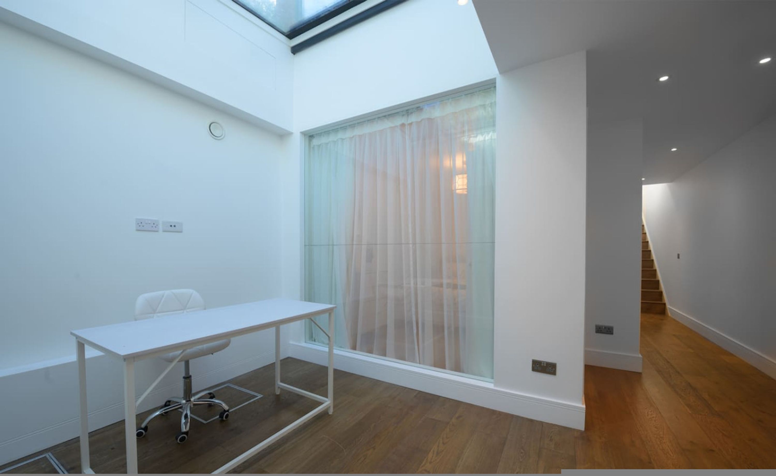 Een moderne, minimalistische kamer met een glazen scheiding, een wit bureau en een trap naar een hoger niveau.