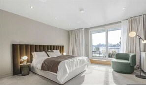 Ein modernes Hotelzimmer mit einem großen Bett, einem Nachttisch, einem grünen Sessel und Blick auf die Stadt durch große Fenster.