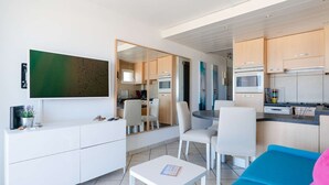 Un salon moderne doté d’un téléviseur à écran plat, d’un coin repas avec des chaises, d’une cuisine équipée de meubles et d’appareils électroménagers, et d’un canapé bleu.