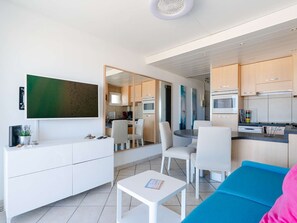 Una sala de estar moderna con televisor de pantalla plana, comedor con sillas, cocina con armarios y electrodomésticos, y un sofá azul.