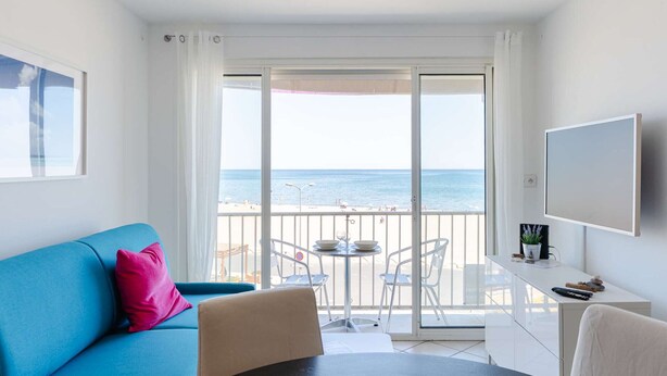 Une pièce avec un canapé bleu, une petite table ronde, un téléviseur et un balcon donnant sur la mer.