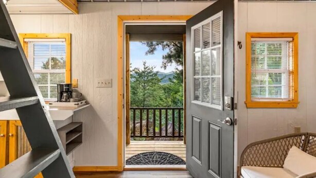 Une pièce dotée d’un escalier en bois, d’une porte donnant sur un balcon avec vue sur les arbres et les montagnes, et d’un fauteuil en osier.