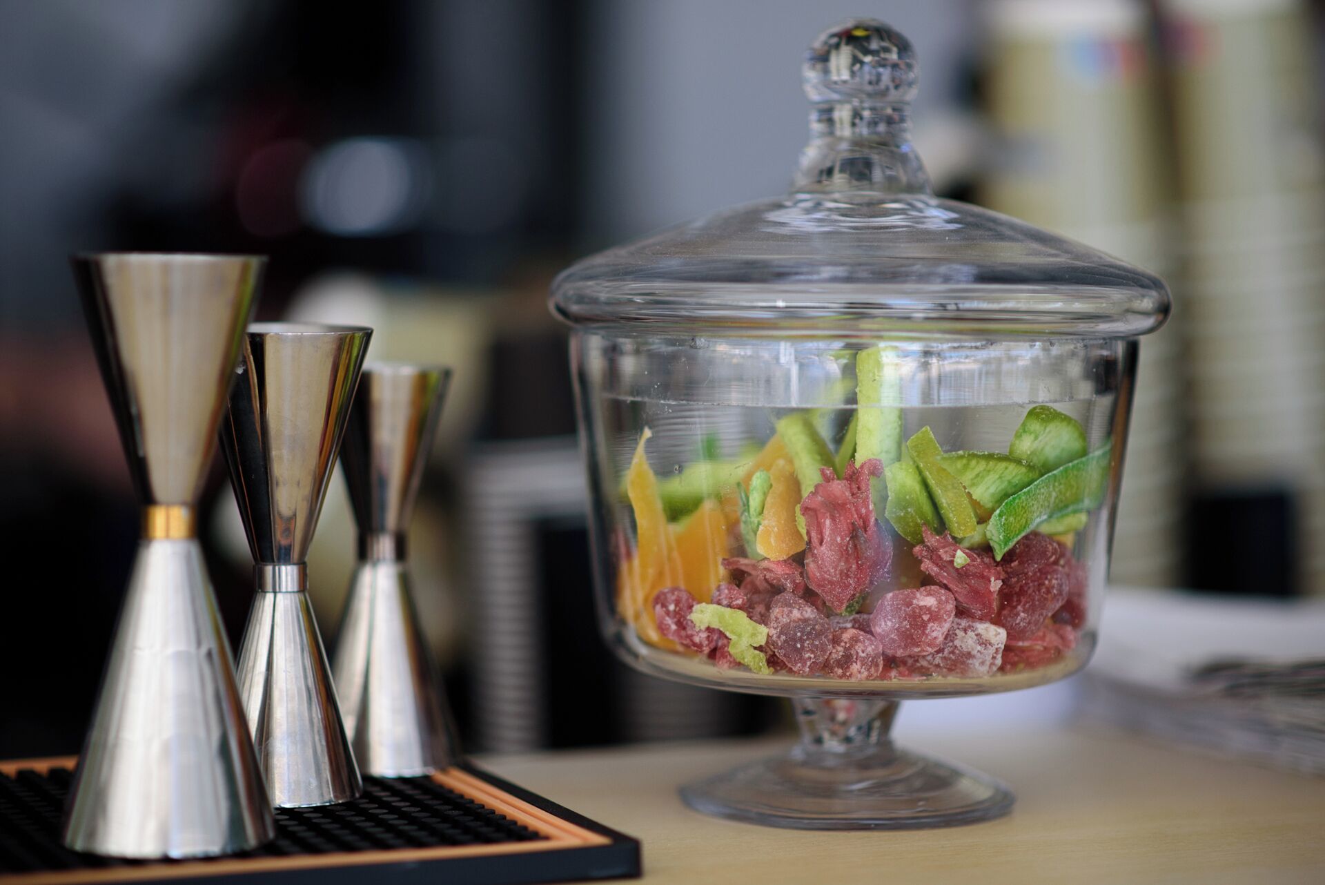 Un recipiente de vidrio transparente con tapa, lleno de frutas y caramelos variados, sobre una mesa con un teclado y un posavasos.