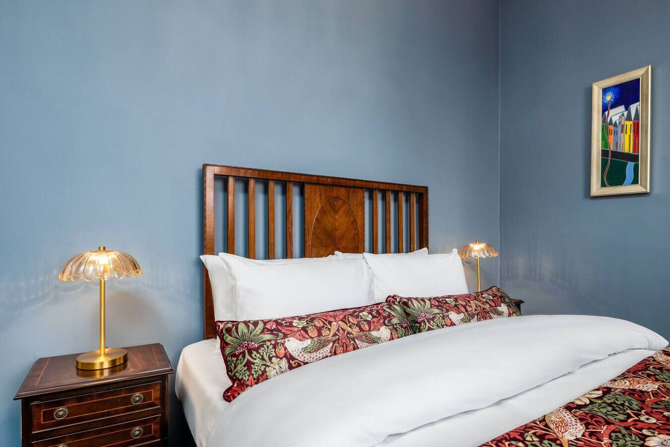 A bedroom with a wooden headboard, a bed with white bedding and floral-patterned pillows, a nightstand with a lamp, and a colorful painting on the wall.