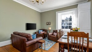 A living room with a brown leather sofa, a wooden dining table with chairs, a chandelier, and a large window with a view. - Elegant 3-Bed Flat | Near Free Parking & Hill View (Stirling)