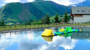 A mountain landscape with a body of water, a wooden house, and a fenced area with colorful floating objects. - Chalet with terrace - 7 guests (Saint-Sorlin-d'Arves)