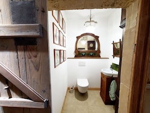 A small, rustic bathroom with a wooden door, a white toilet, a wooden vanity with a mirror, and framed pictures on the wall.