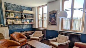 A cozy living room with leather sofas, a wooden coffee table, and a patterned rug. There are bookshelves with books and decorative items, and a large window with curtains.