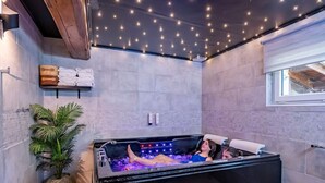 Une salle de spa moderne avec un grand jacuzzi, une plante en pot et une étagère avec des serviettes.