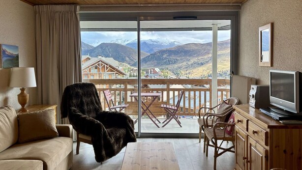 Una sala de estar acogedora con sofá, sillón y mesa de centro, un televisor y un balcón con vistas a las montañas.