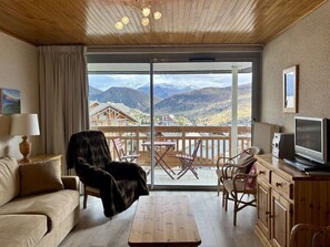 A cozy living room with a sofa, armchair, and coffee table, a TV, and a balcony with a view of mountains. - Convenient studio, 5 guests, near center (83 units) (Huez)