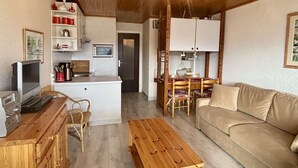 Una sala de estar acogedora con techo de madera, un sofá, una mesa de centro de madera, un comedor con mesa y sillas, y una kitchenette con armarios y un microondas.