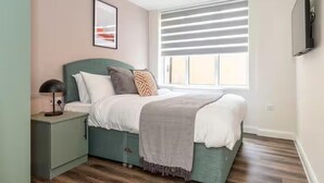 Een slaapkamer met een bed, nachtkastje, lamp, raam met zonwering en een lijst met een afbeelding aan de muur.