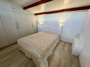 Quarto com cama, piso de madeira, paredes brancas e teto com vigas aparentes.