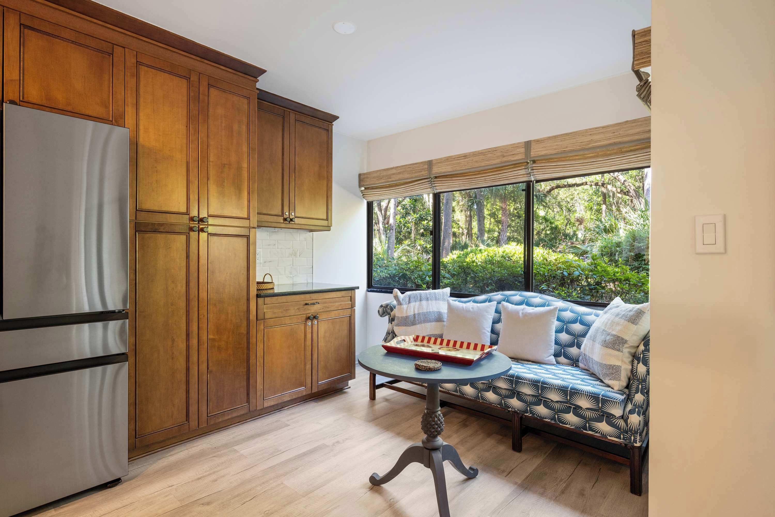A kitchen with wooden cabinets, a stainless steel refrigerator, a dining table with a tray, and a patterned sofa by a window.