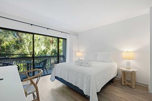 Habitación de hotel con cama, dos lámparas de noche, una mesita de noche de madera, una silla y un balcón con vistas a zonas verdes.