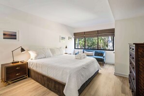 Ein Schlafzimmer mit einem großen Bett, einem Nachttisch, einer Kommode, einem Fenster mit Jalousien und Blick auf Bäume.