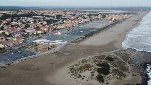 Luftaufnahme einer Küstenstadt mit breitem Strand, einem großen Platz und einem Gebäude mit markanter Dachkonstruktion.