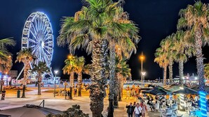 Ein beleuchteter Außenbereich bei Nacht mit einem großen Riesenrad, Palmen und Spaziergängern.