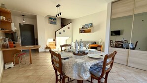 On aperçoit un coin repas avec une table dressée pour quatre personnes, entourée de chaises en bois. Une cuisine est visible sur la gauche, avec un réfrigérateur et un évier. Un escalier mène à l’étage supérieur.