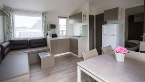 A compact kitchenette with a microwave, stove, and refrigerator. There is a seating area with a sofa and a small table. A window provides natural light. - Mobile home with terrace - 6 guests (Binic)