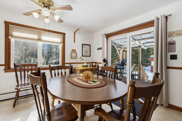 A dining area with a round wooden table, wooden chairs, a ceiling fan, and large windows with a view of trees.