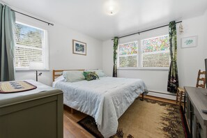 Un dormitorio con una cama, un escritorio y una silla. Hay dos ventanas con persianas, una lámpara de techo y un cuadro en la pared.