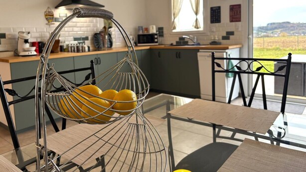 A modern kitchen with a hanging lemon basket, a dining area with a glass table, and a view of the countryside.