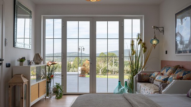 Ein Schlafzimmer mit großem Fenster, einem Bett, einem Schminktisch und Blick ins Freie.