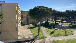 Un edifici de diversos pisos amb balcons, un passeig pavimentat i una zona verda amb arbres.