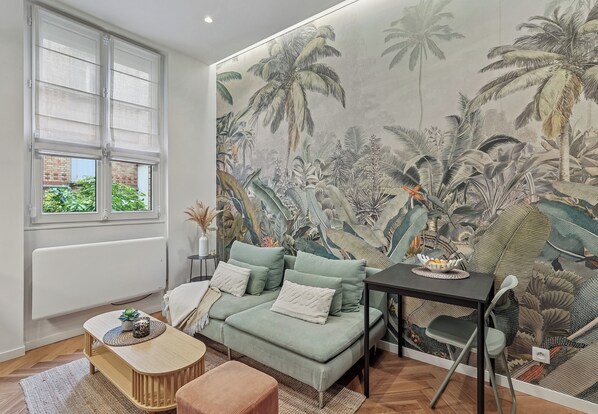 Ein Wohnzimmer mit einem grünen Sofa, einem kleinen Holztisch und einer Wandmalerei von tropischen Pflanzen.