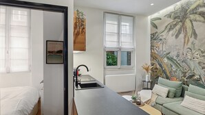 Un espacio habitable compacto con kitchenette, una cama, un sofá verde y un mural tropical grande.