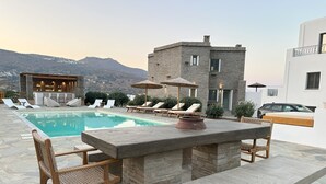 A poolside area with a stone table and chairs, lounge chairs, and a bar area with a mountain view in the background. - Ammos Andros Accommodation (Ormos Korthiou)