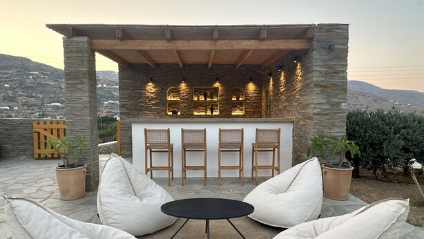 An outdoor seating area with stone walls, a bar, and a view of the mountains. - Ammos Andros Accommodation (Ormos Korthiou)