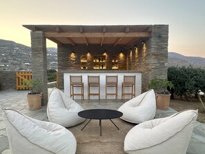 An outdoor seating area with stone walls, a bar, and a view of the mountains. - Ammos Andros Accommodation (Ormos Korthiou)