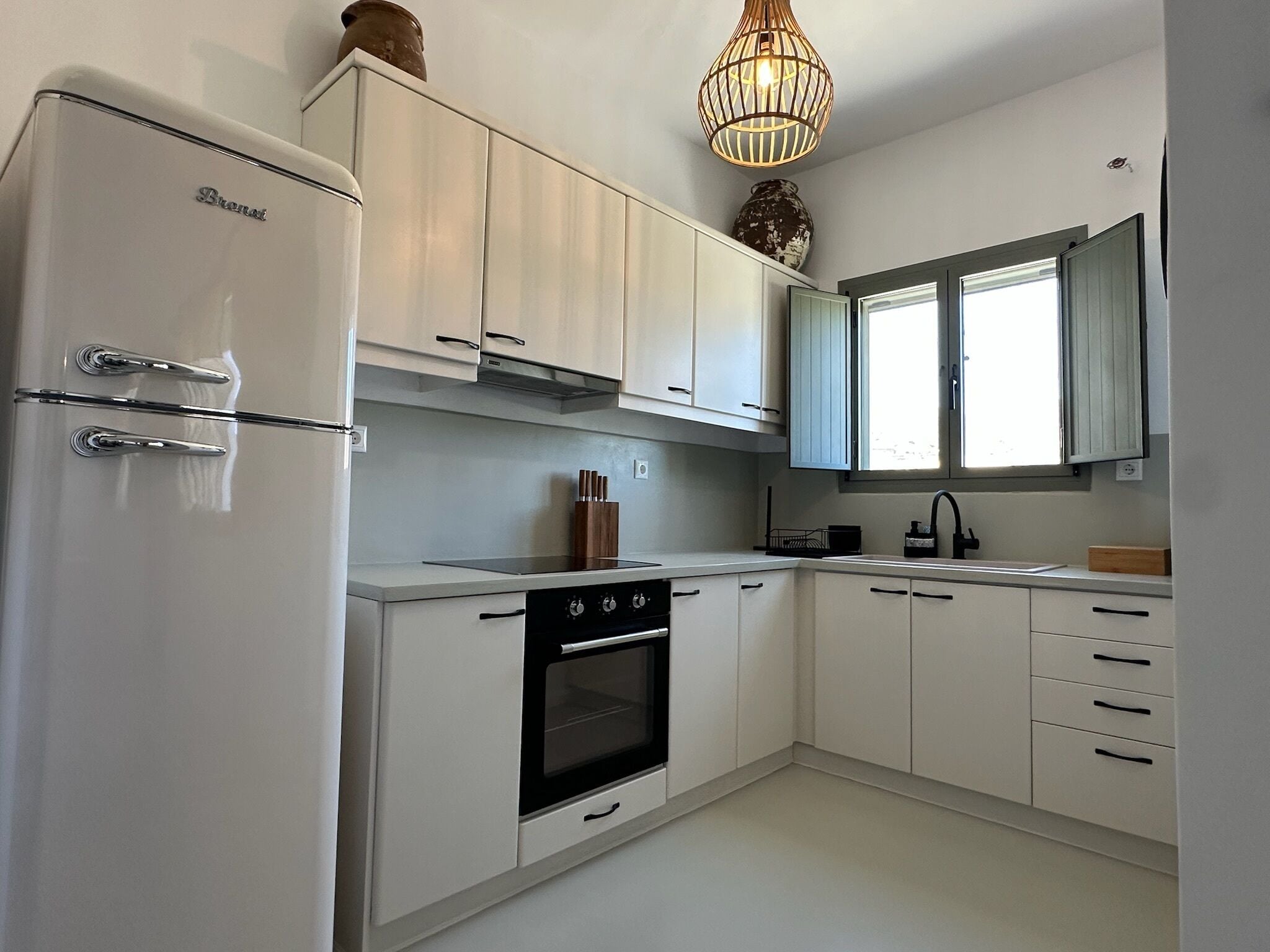 A modern kitchen with white cabinetry, a built-in oven, a refrigerator, and a window with blinds.