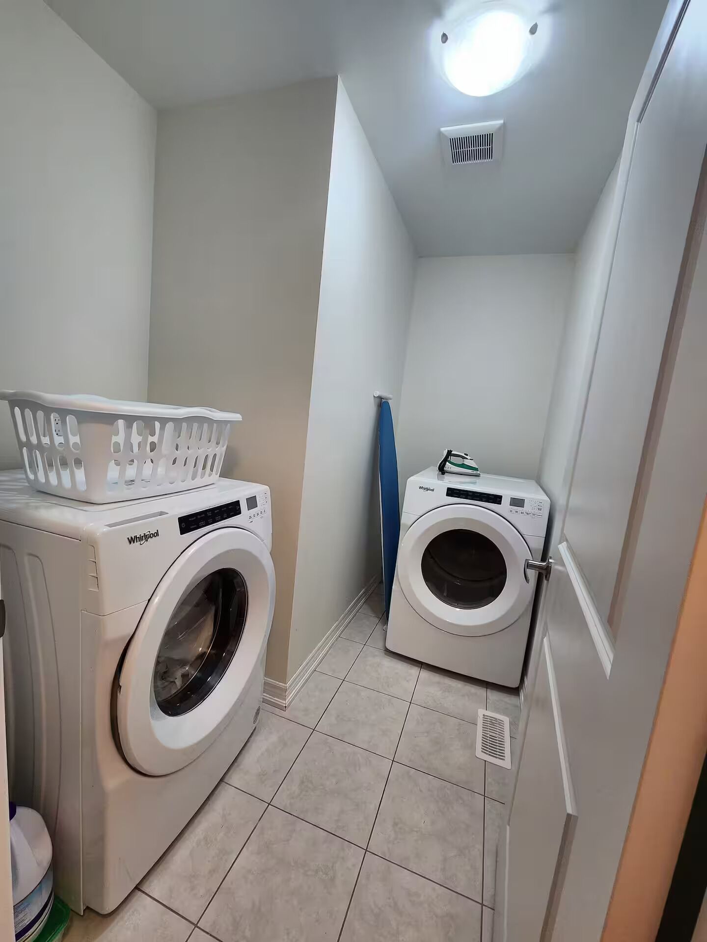 A compact laundry room with two Whirlpool washing machines, a white laundry basket, and a blue ironing board.