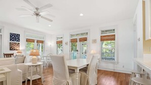 Un intérieur lumineux et blanc, avec un coin repas comprenant une table et des chaises blanches, un ventilateur de plafond et de grandes fenêtres équipées de stores.