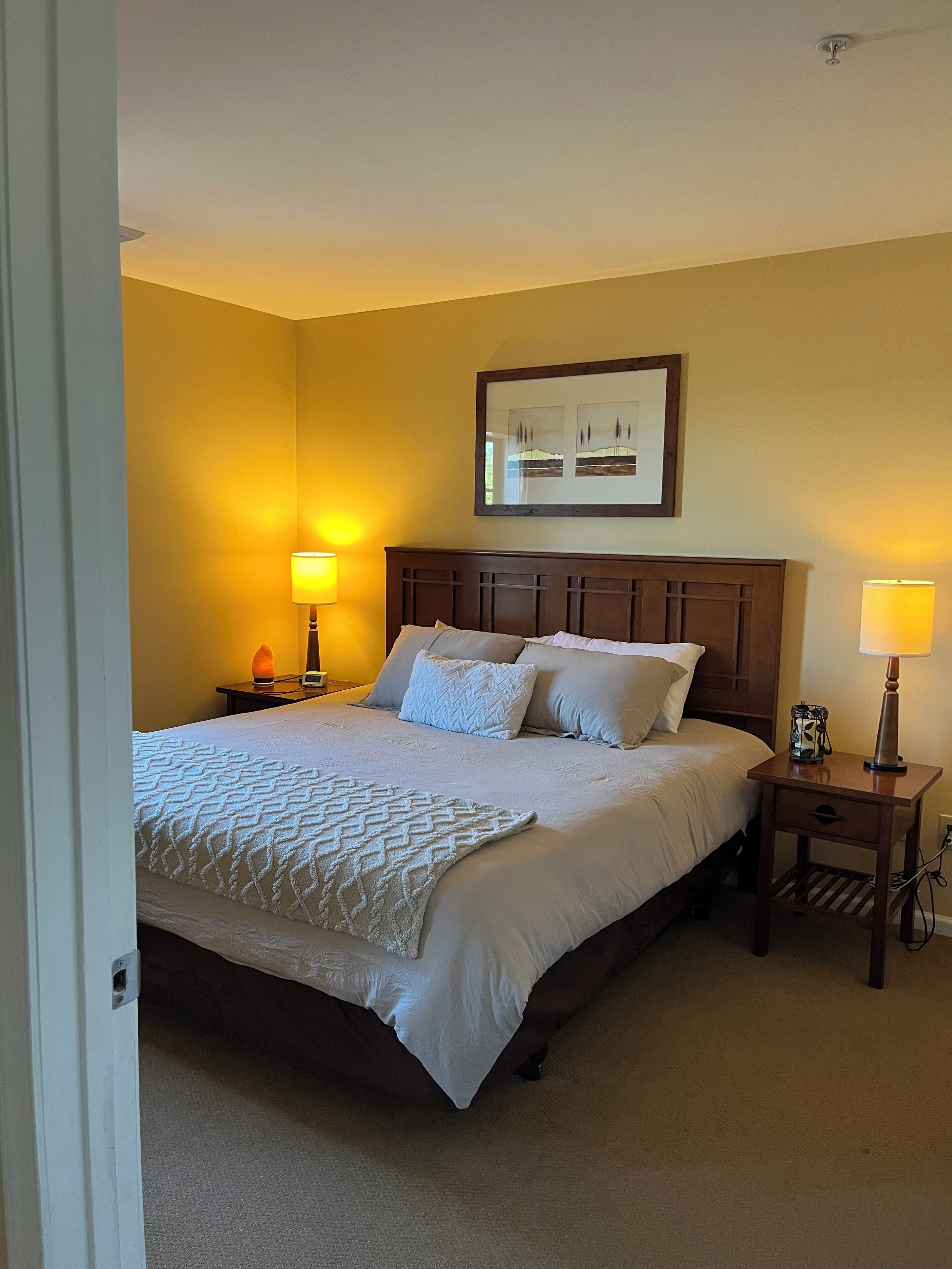 A neatly made bed with a white comforter and pillows, two bedside lamps, a framed picture on the wall, and a wooden nightstand with a lamp.